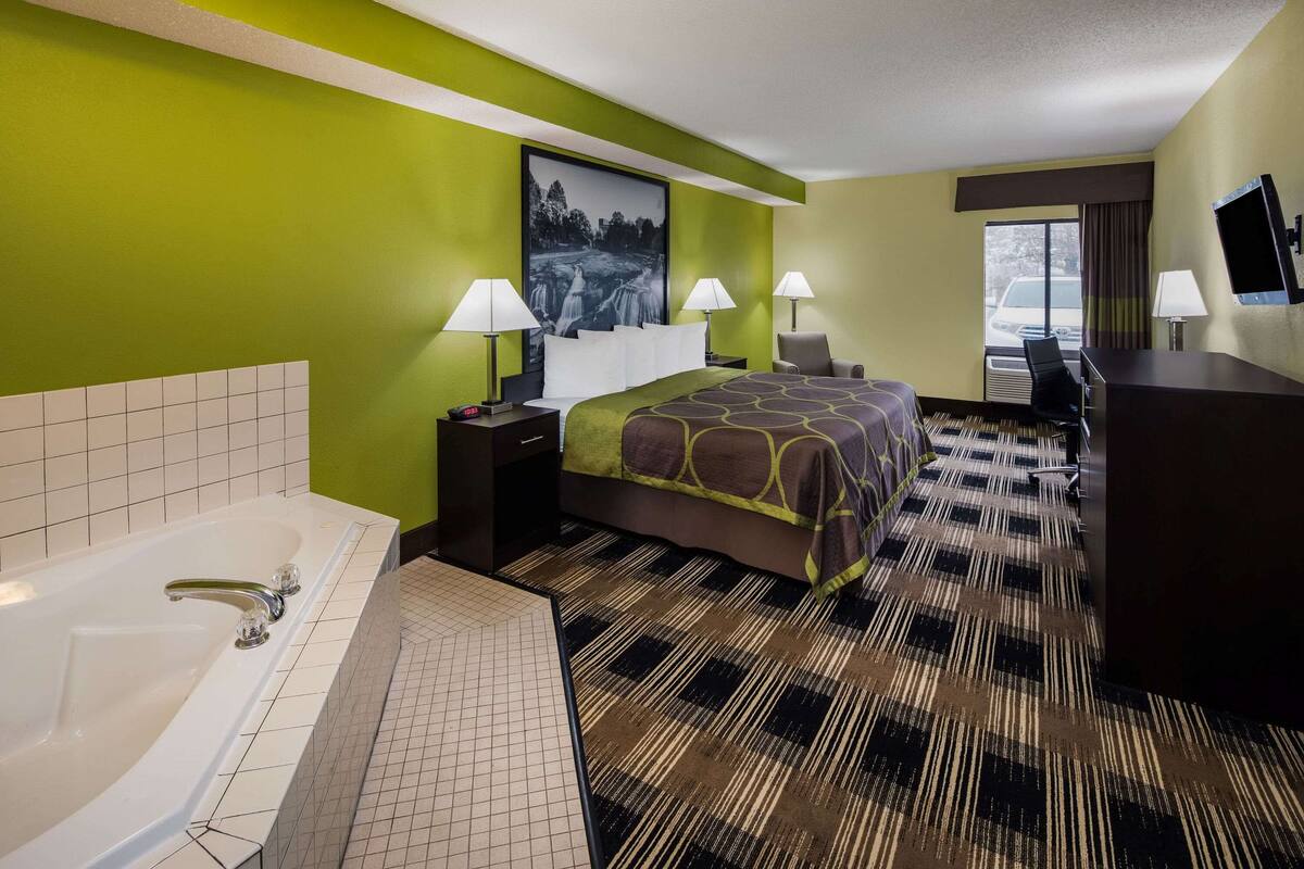 Une chambre d’hôtel avec un grand lit, deux lampes de chevet, un bureau avec une chaise, une télévision et une salle de bain avec une baignoire et des toilettes.