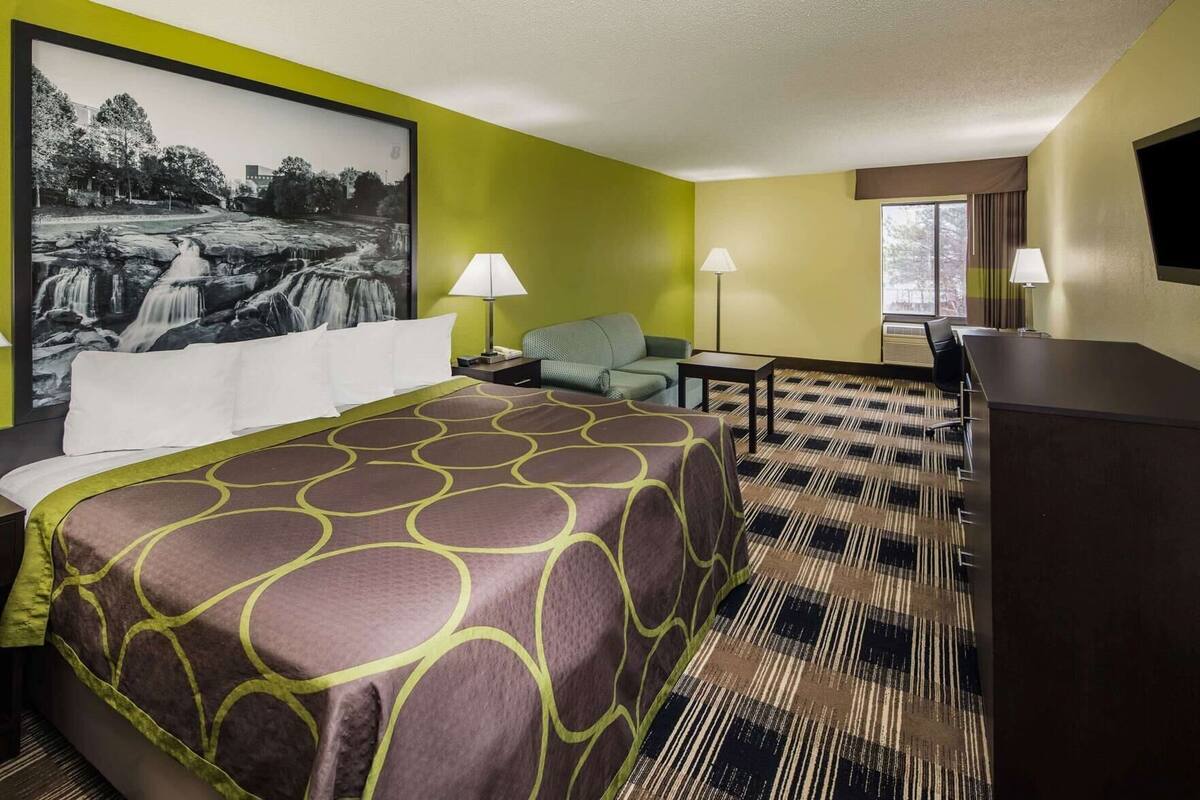 Une chambre d’hôtel avec un lit, un bureau, une chaise, deux lampes et un grand tableau encadré au mur.