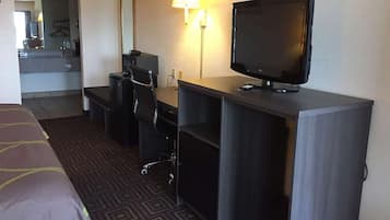 A hotel room with a bed, a desk with a computer and a TV, a chair, and a lamp.