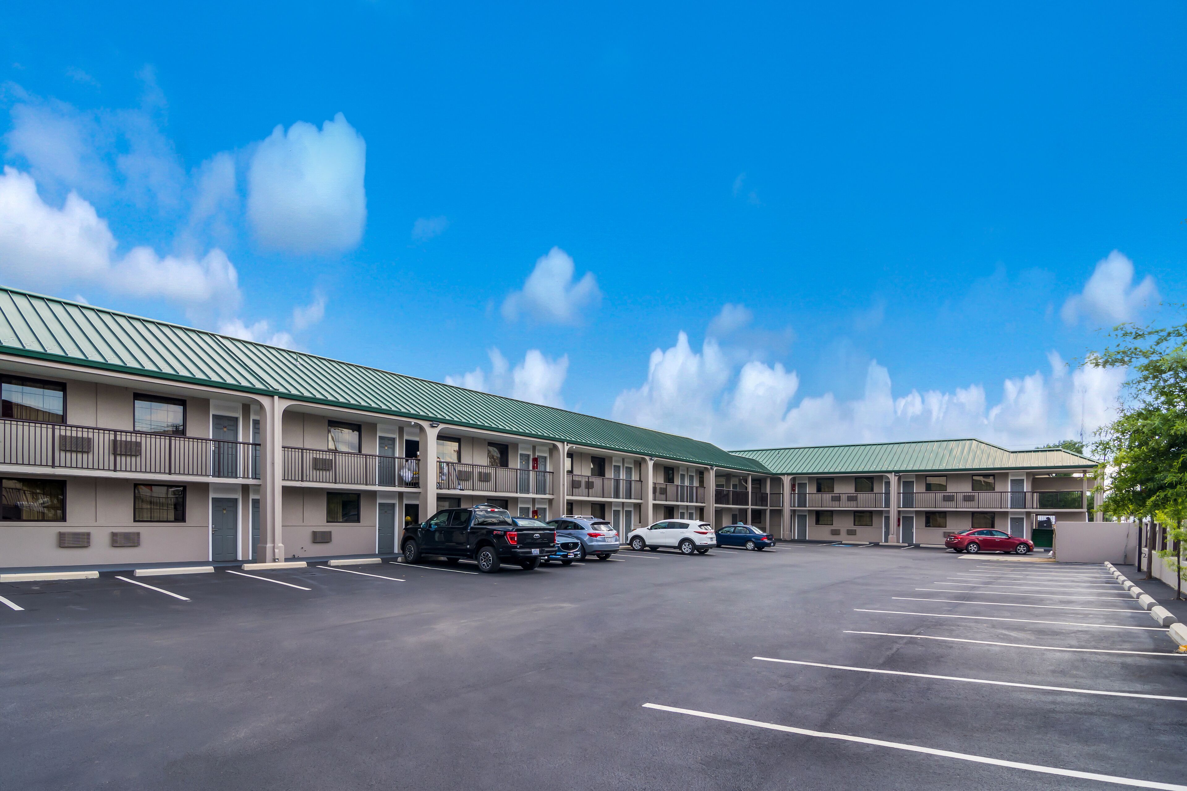 A motel with a green roof, multiple floors, and a parking lot with several cars.