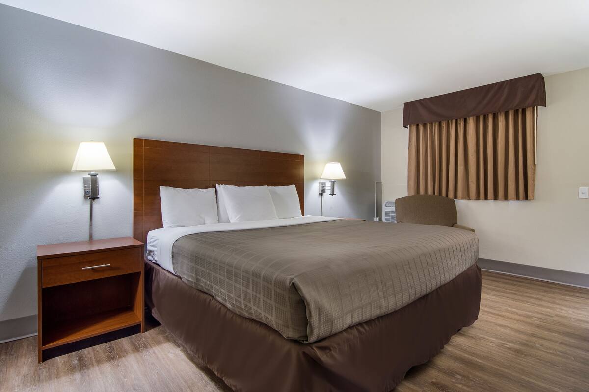 A hotel room with a wooden headboard, a bed with a brown bedspread, two bedside lamps, a nightstand, and a window with curtains.