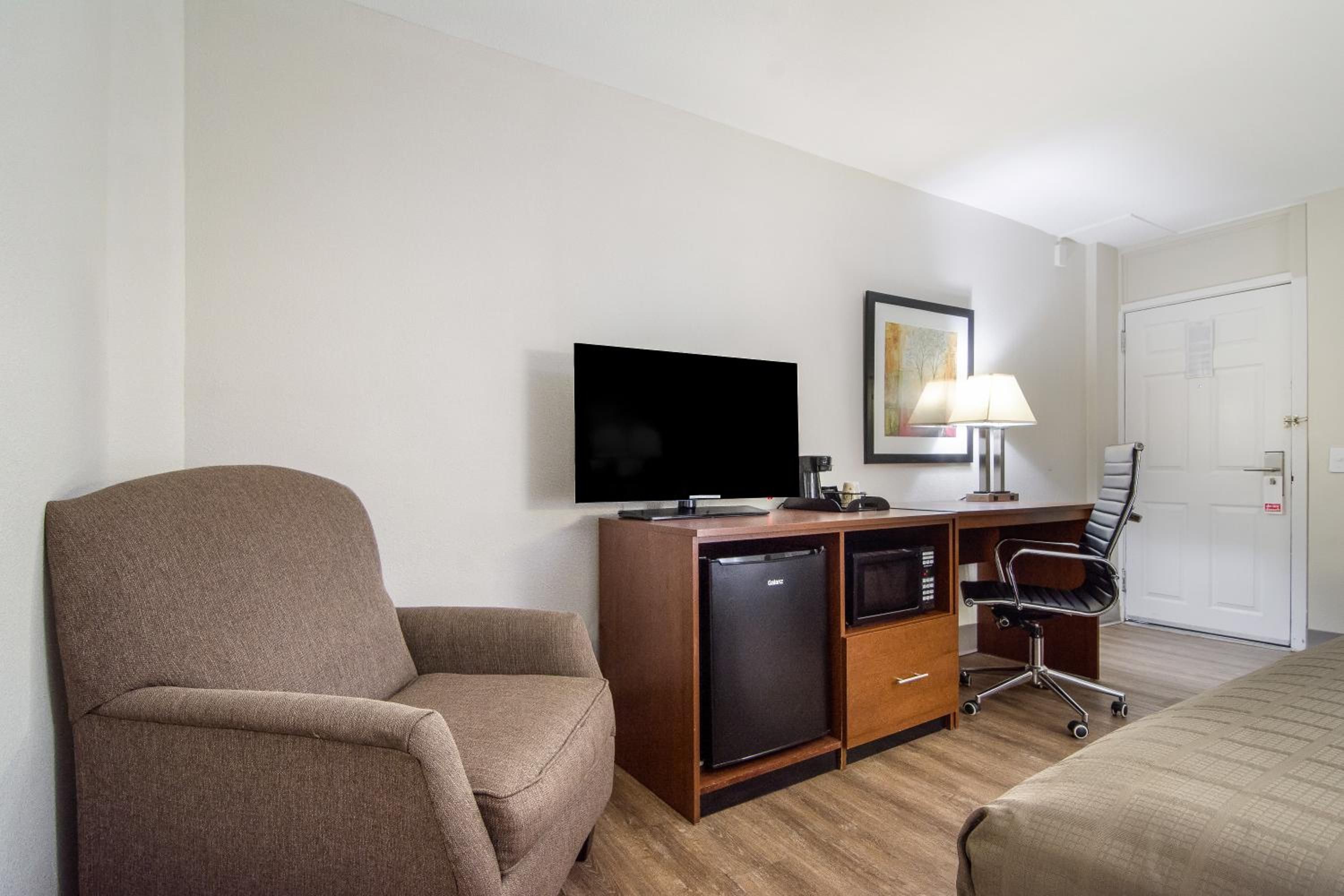 A hotel room with a single bed, a desk with a computer, a television, a chair, and a small refrigerator.
