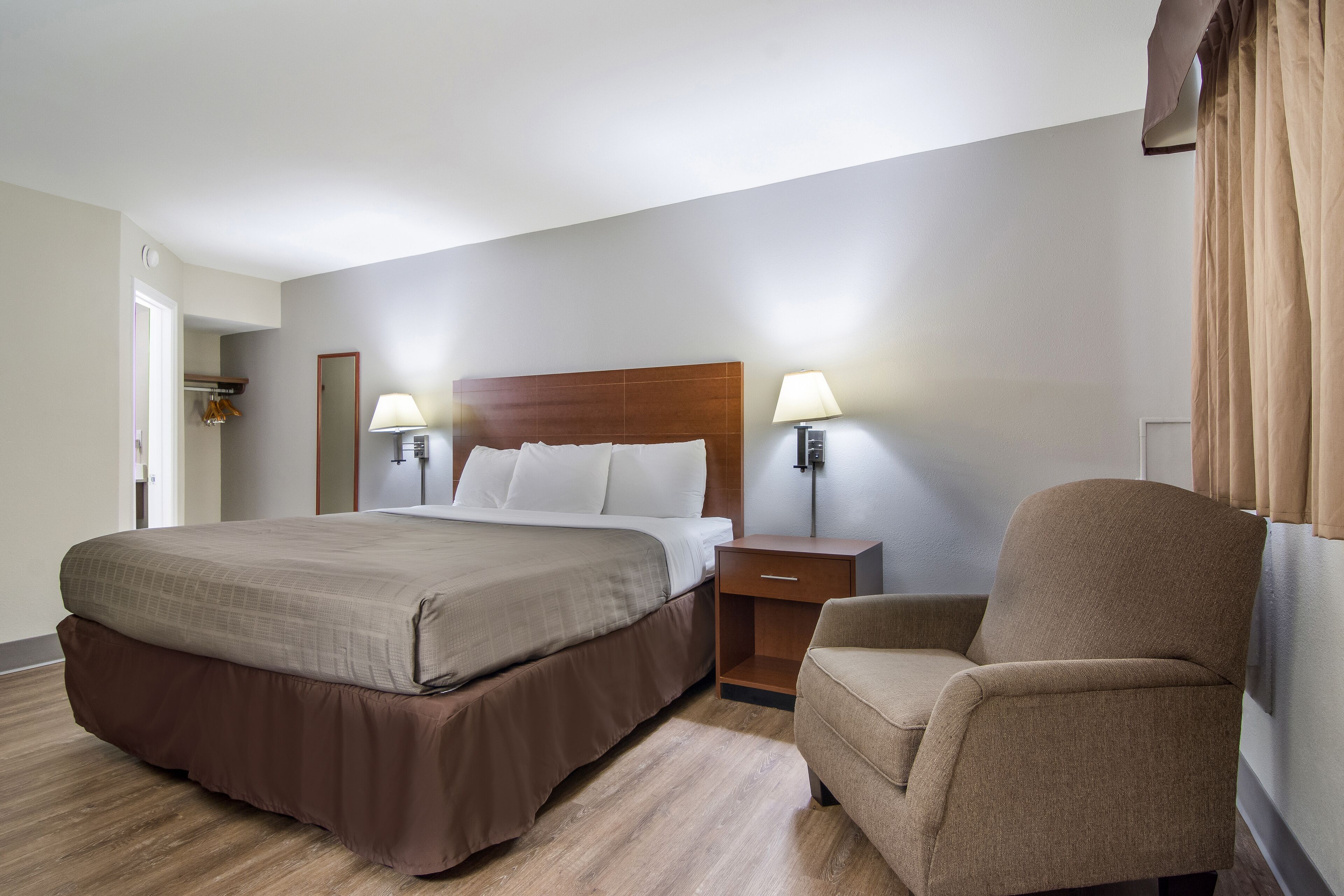Een hotelkamer met een groot bed, twee bedlampen, een houten nachtkastje, een stoel en een deur naar een inloopkast.