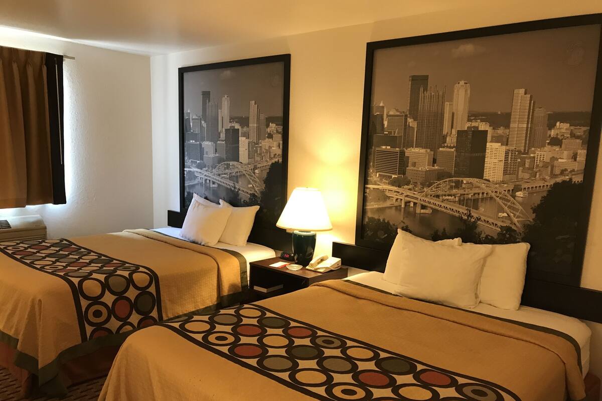Habitación de hotel con dos camas, una mesita de noche con lámpara y un cuadro grande con un paisaje urbano en la pared.