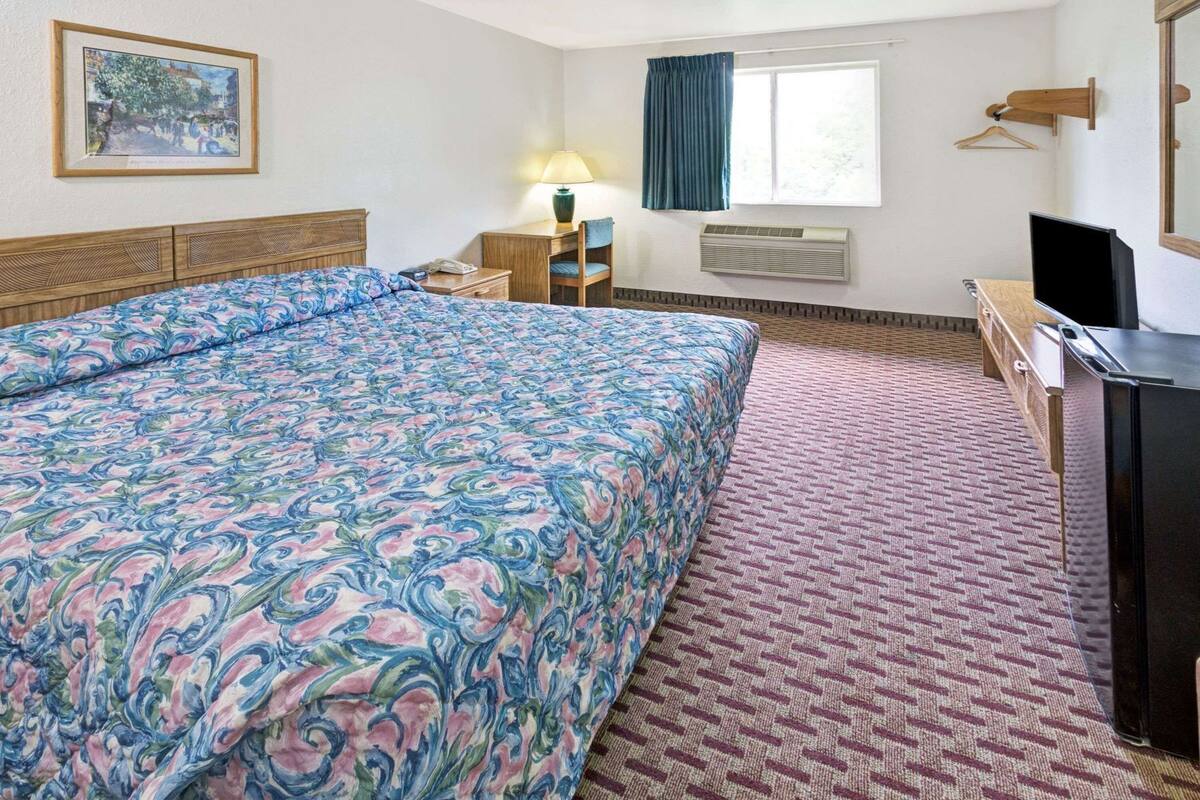 Habitación de hotel con una cama grande, un escritorio con computadora, una ventana con cortinas, aire acondicionado y un cuadro en la pared.