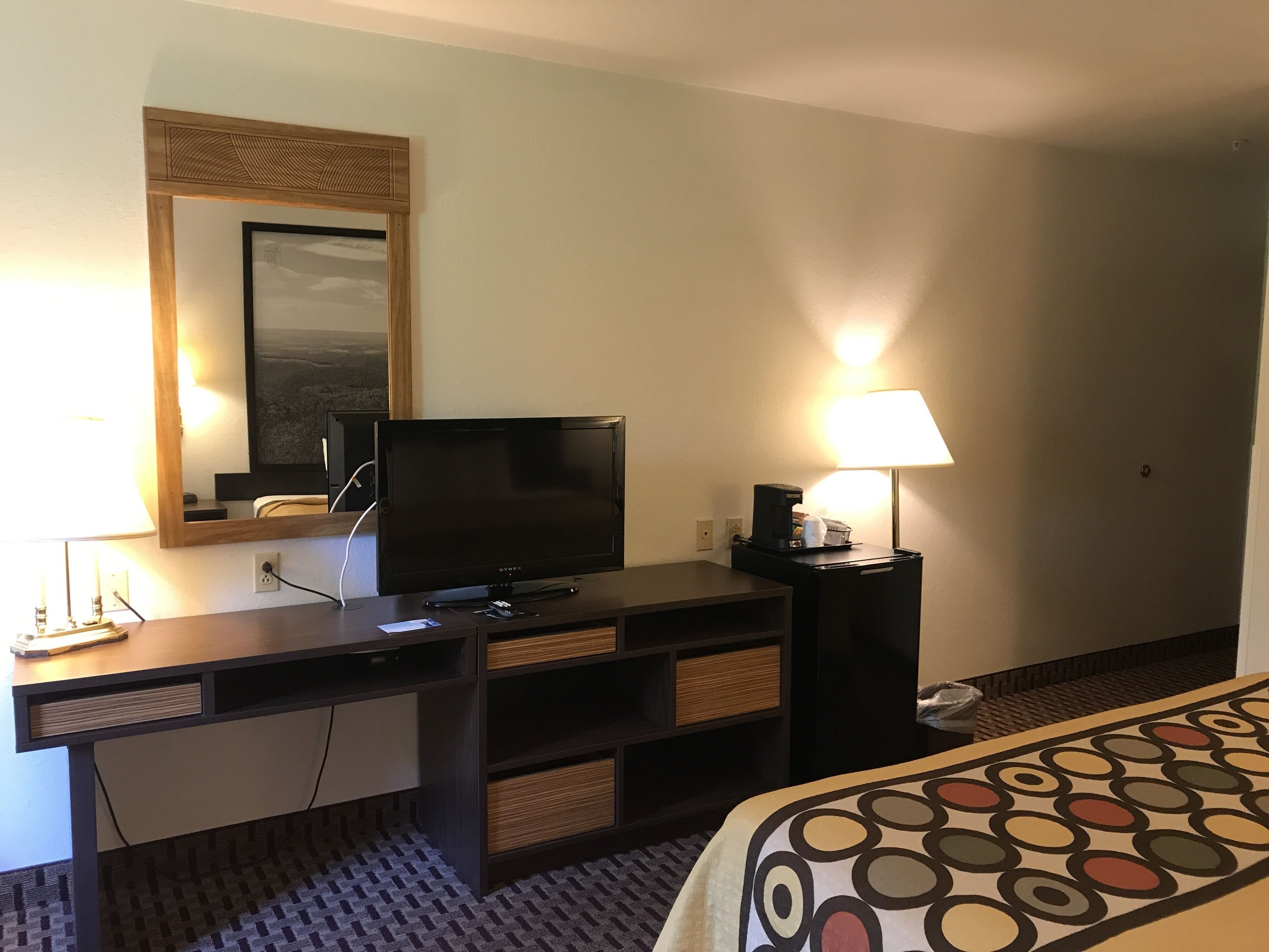 A hotel room with a desk, a television, a lamp, and a bed with a patterned bedspread.