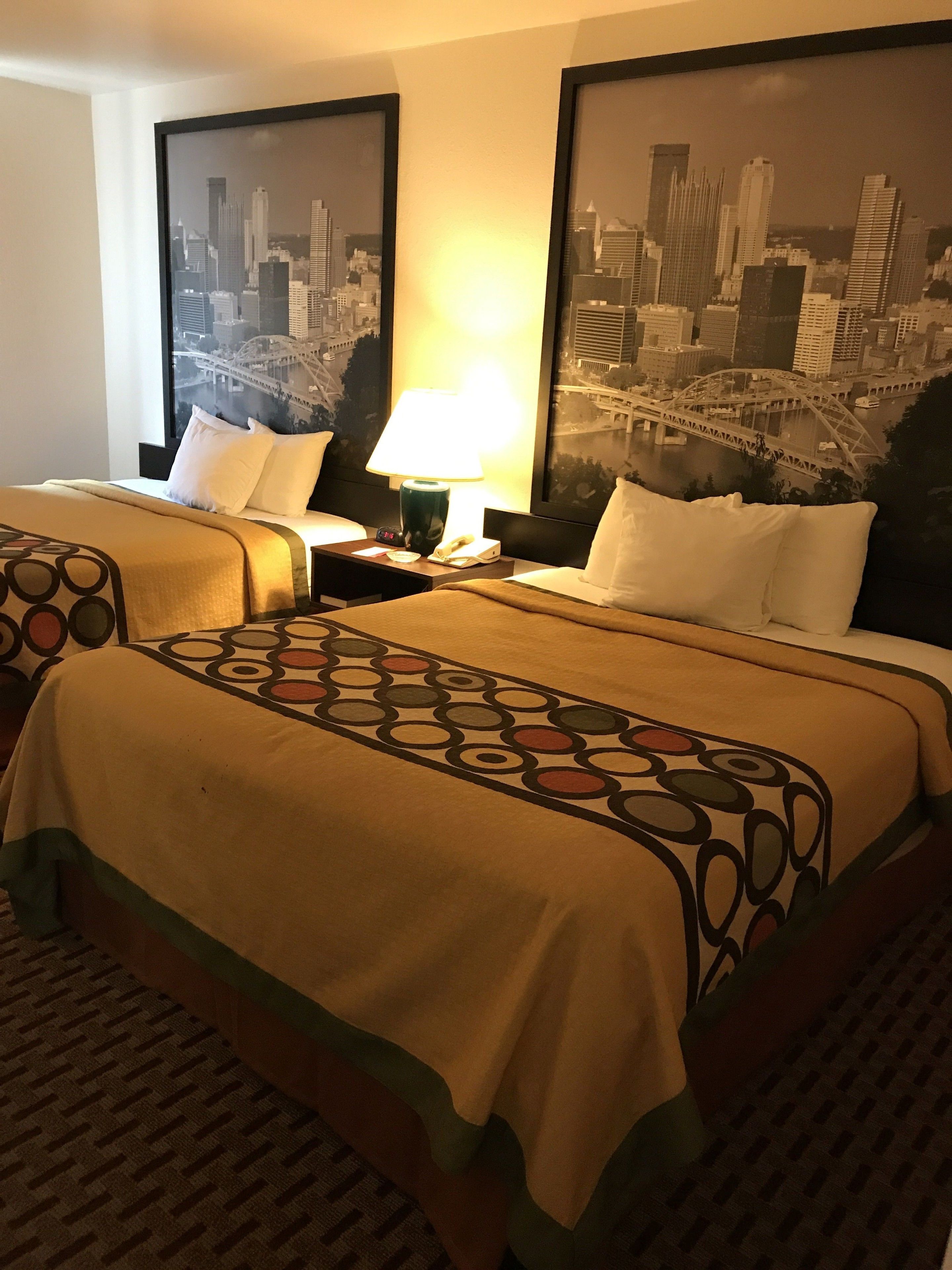 A hotel room with two beds, a nightstand with a lamp, and two framed cityscape pictures on the wall.