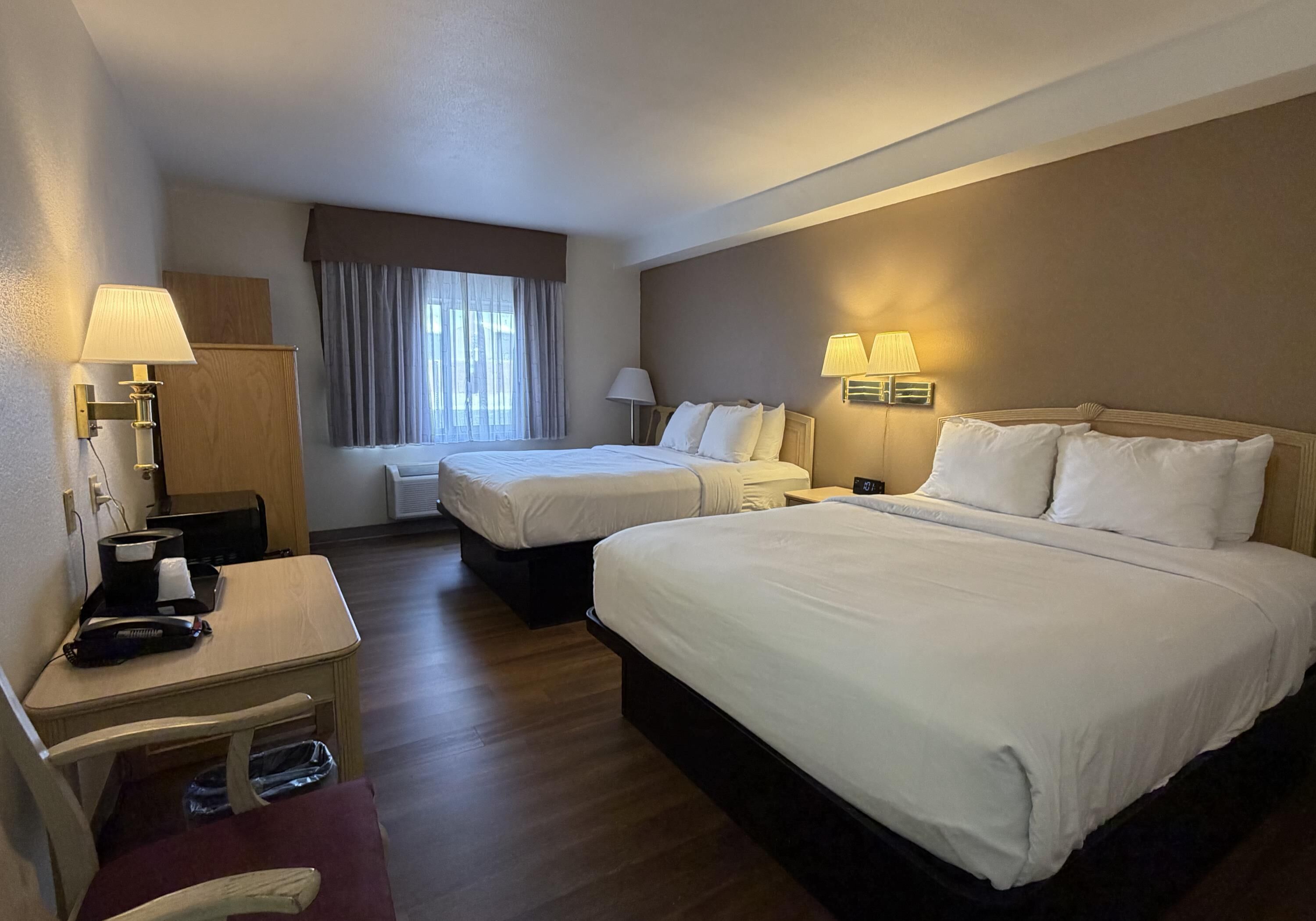 A hotel room with two beds, a desk, a chair, a lamp, and a window with curtains.