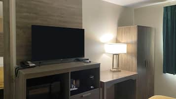 A hotel room with a flat-screen TV on a wooden entertainment unit, a desk with a lamp, a wardrobe, and a bed with a yellow cover.