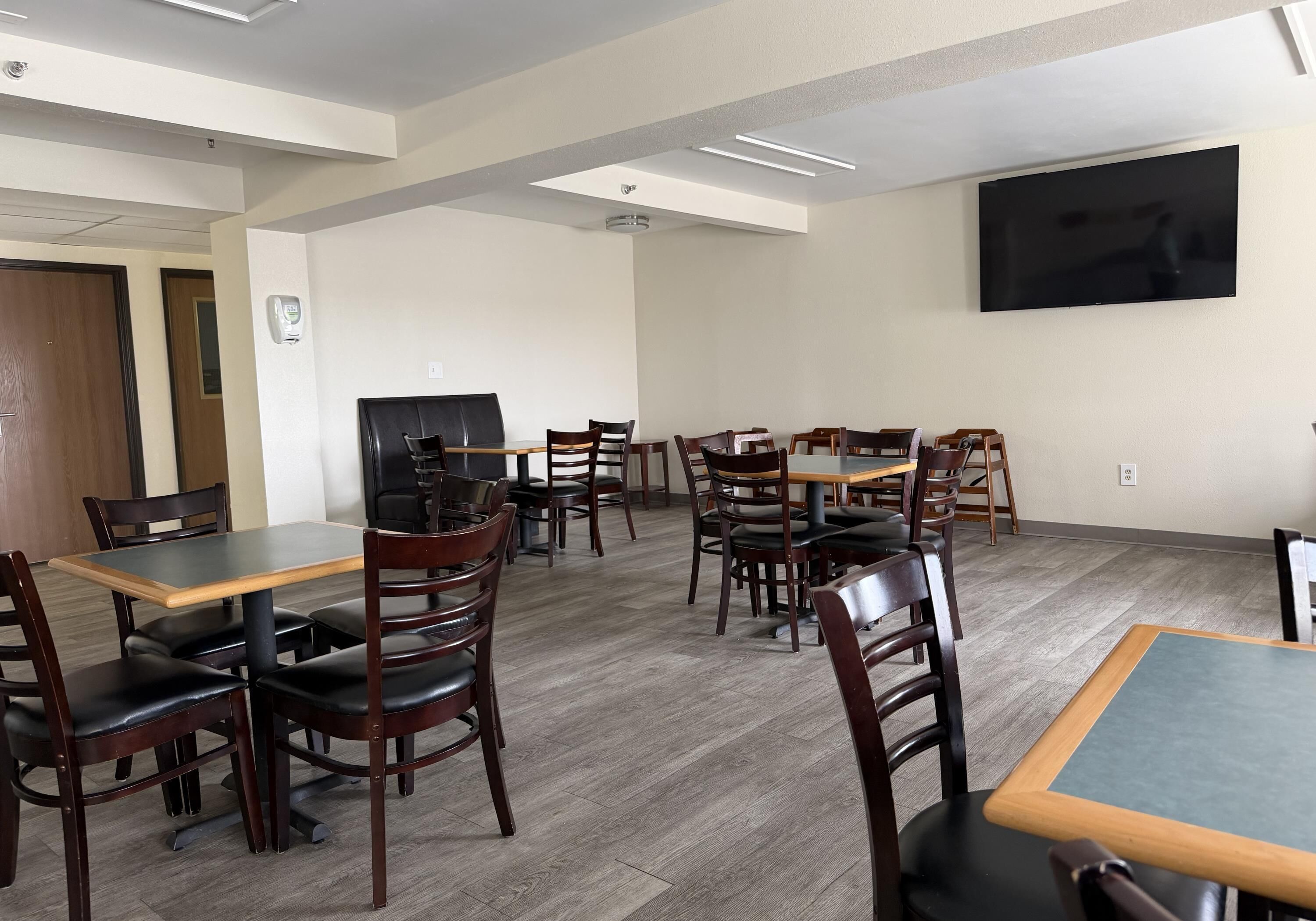 A hotel dining area with multiple tables and chairs, a flat-screen TV mounted on the wall, and a ceiling with recessed lighting.