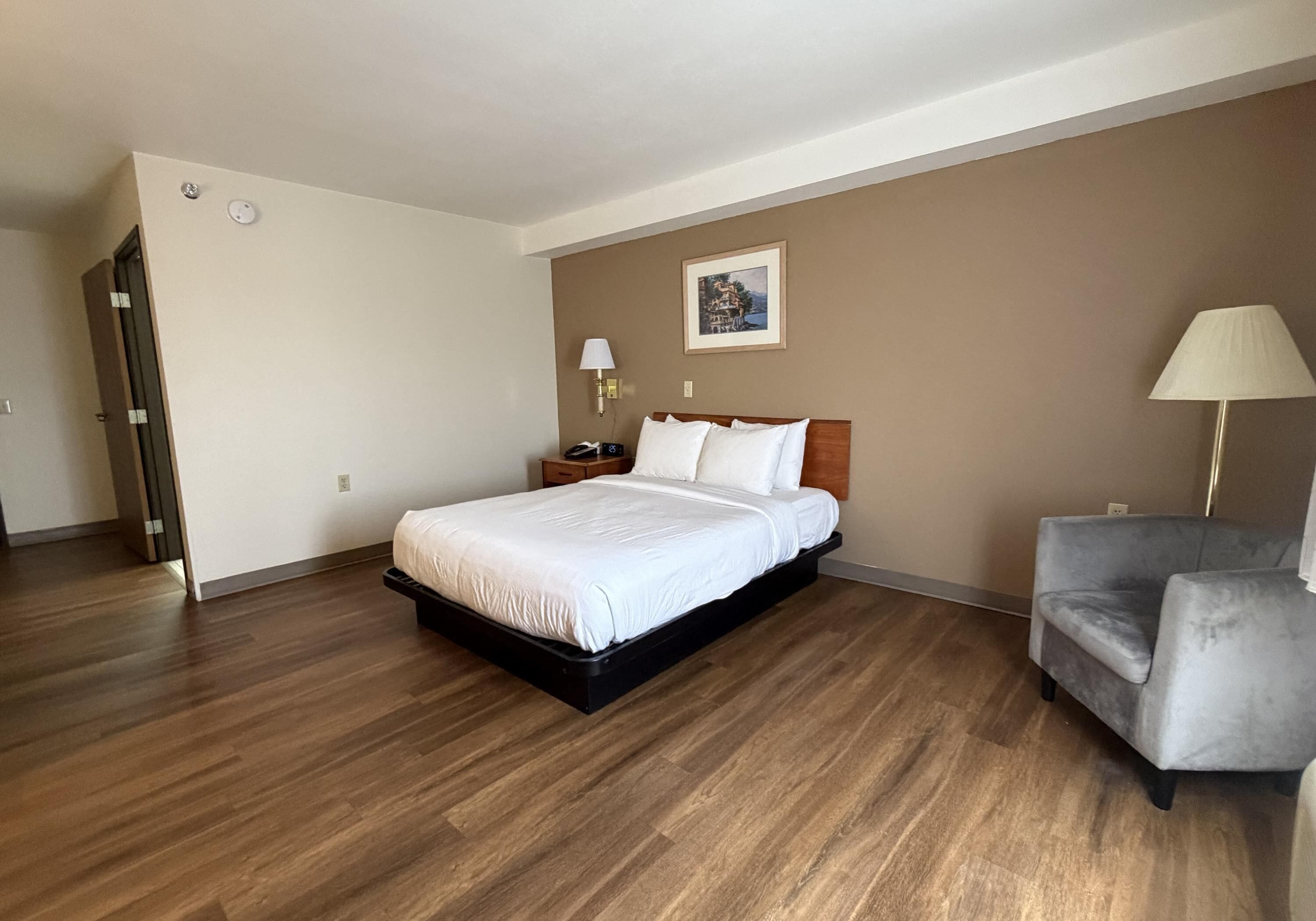 A hotel room with a bed, a chair, a nightstand, a lamp, and a painting on the wall.