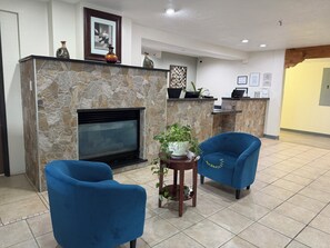 Un hall con chimenea de piedra, dos sillones azules, una mesita con una planta y un mostrador de recepción con computadoras portátiles.