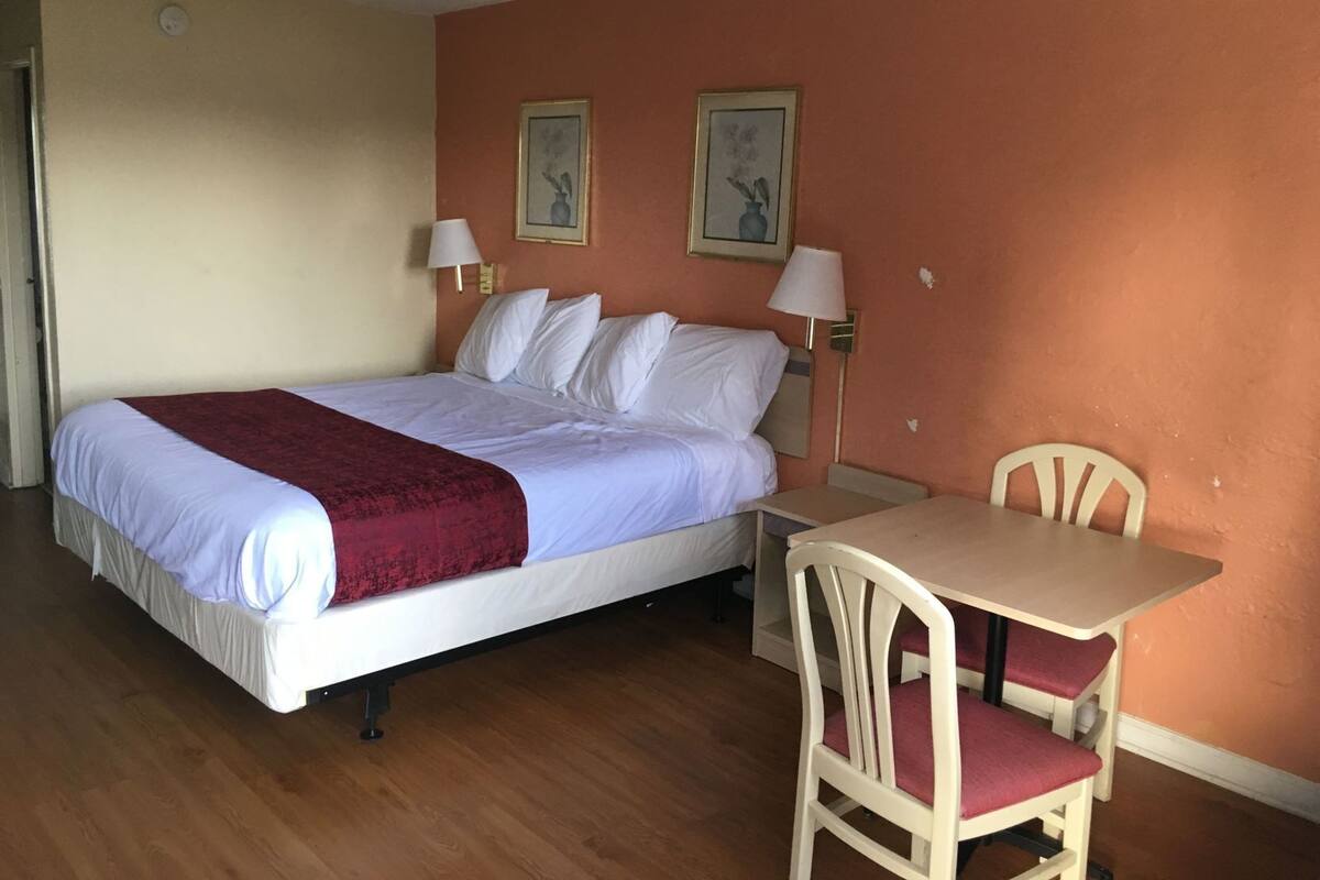 A hotel room with a bed, a table, a chair, two wall-mounted lamps, and two framed pictures.