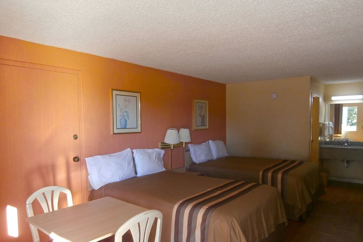 A hotel room with two beds, a table, a chair, and a bathroom with a mirror and sink.