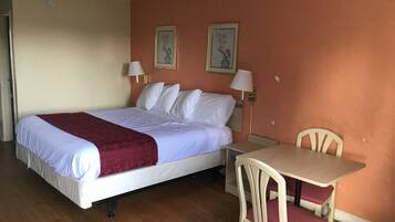 Uma cama de solteiro com um cobertor vermelho, duas luminárias fixadas na parede, uma pequena mesa com uma cadeira e duas fotografias emolduradas na parede.
