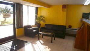 A room with a yellow wall, a black leather sofa, a black armchair, a small table with magazines, a potted plant, a clock, and a sign indicating a night window. - Americas Best Value Inn Tucker (Tucker)