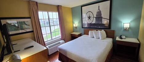 A hotel room with a bed, a desk, a chair, a window with curtains, a wall-mounted TV, a wall-mounted lamp, and a framed picture of a building.