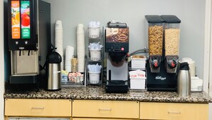 Una estación de desayuno autoservicio con dispensadores de bebidas, una máquina de café y recipientes para cubiertos y cereales.