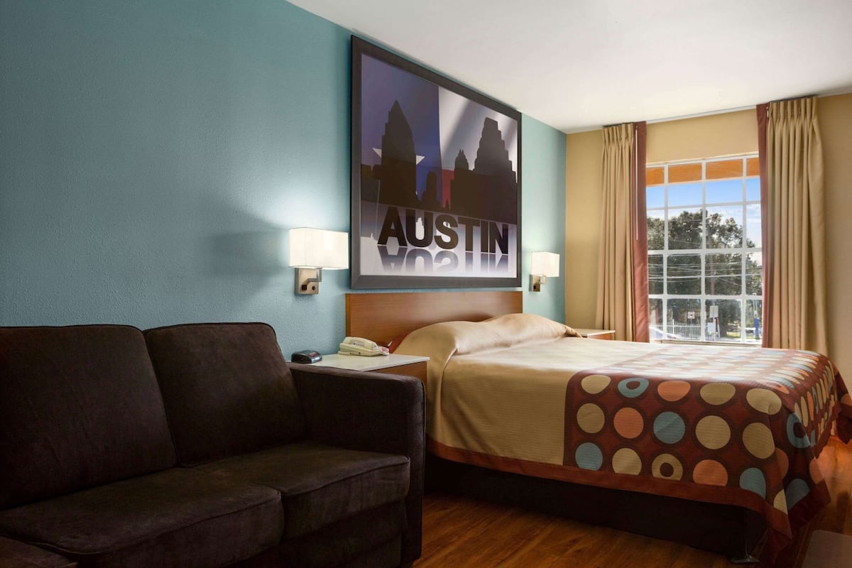 Una camera d'albergo con un letto, una poltrona, una TV a parete e una finestra con tende.