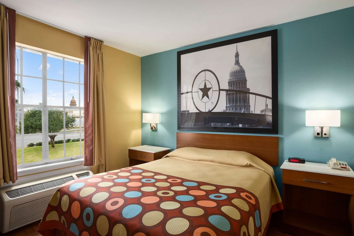 Una camera d'albergo con un letto, una finestra con tende, una lampada a parete, un quadro incorniciato che raffigura un edificio e un comodino con un telefono e una sveglia.