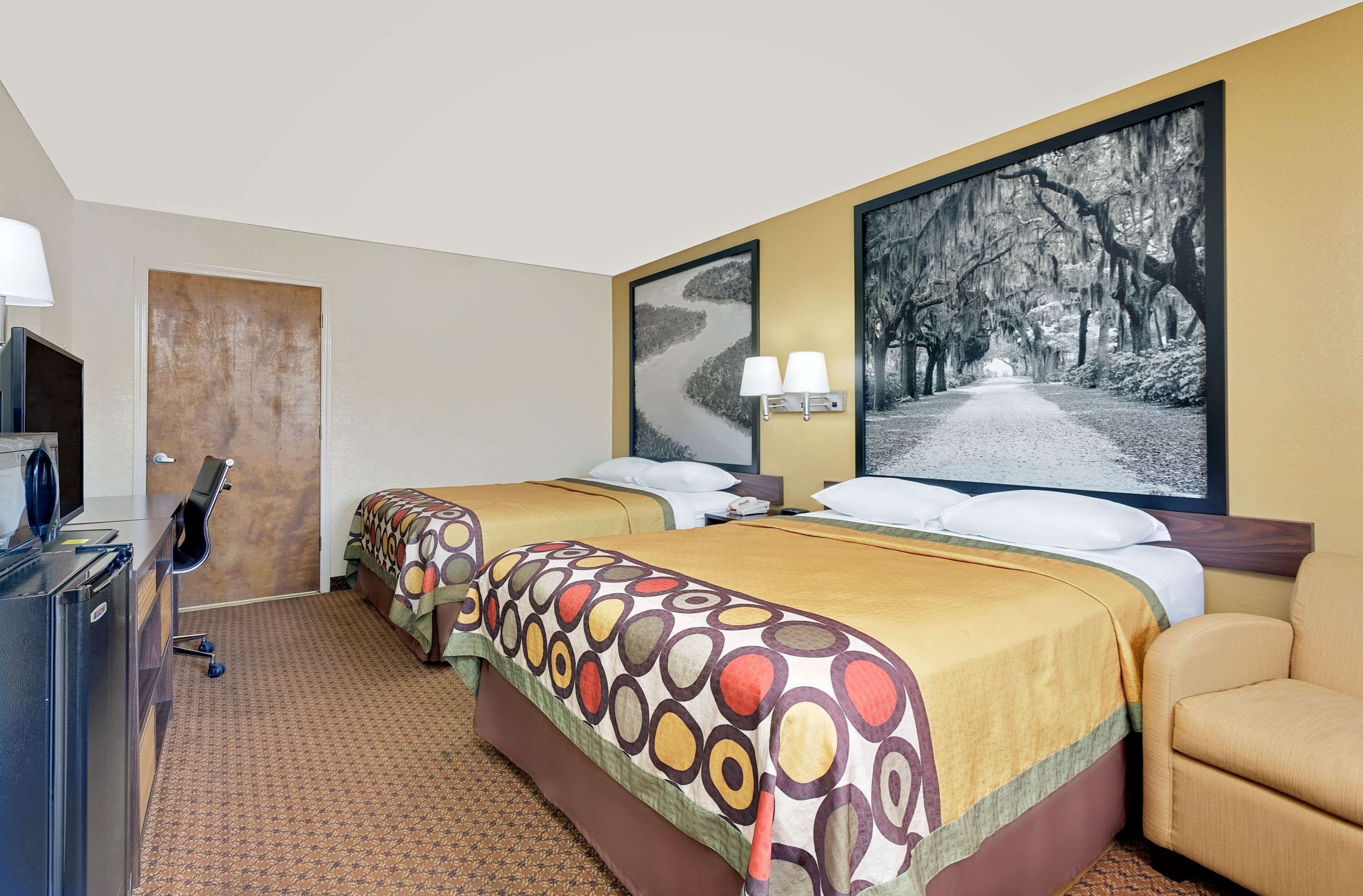 A hotel room with two beds, a desk with a lamp, and a large framed picture of a tree-lined road.