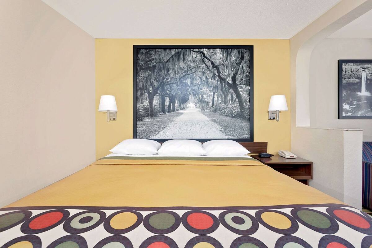 Hotelkamer met een bed, twee wandlampen, een foto in een lijst boven het bed, en een klein nachtkastje met een telefoon.
