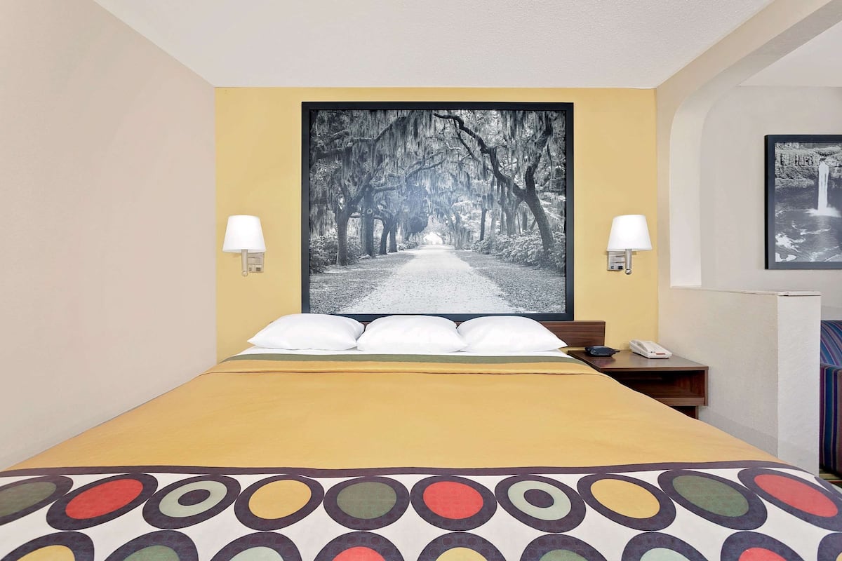 Een hotelkamer met een bed, nachtkastje, lamp en een groot schilderij van een weg omzoomd met bomen.