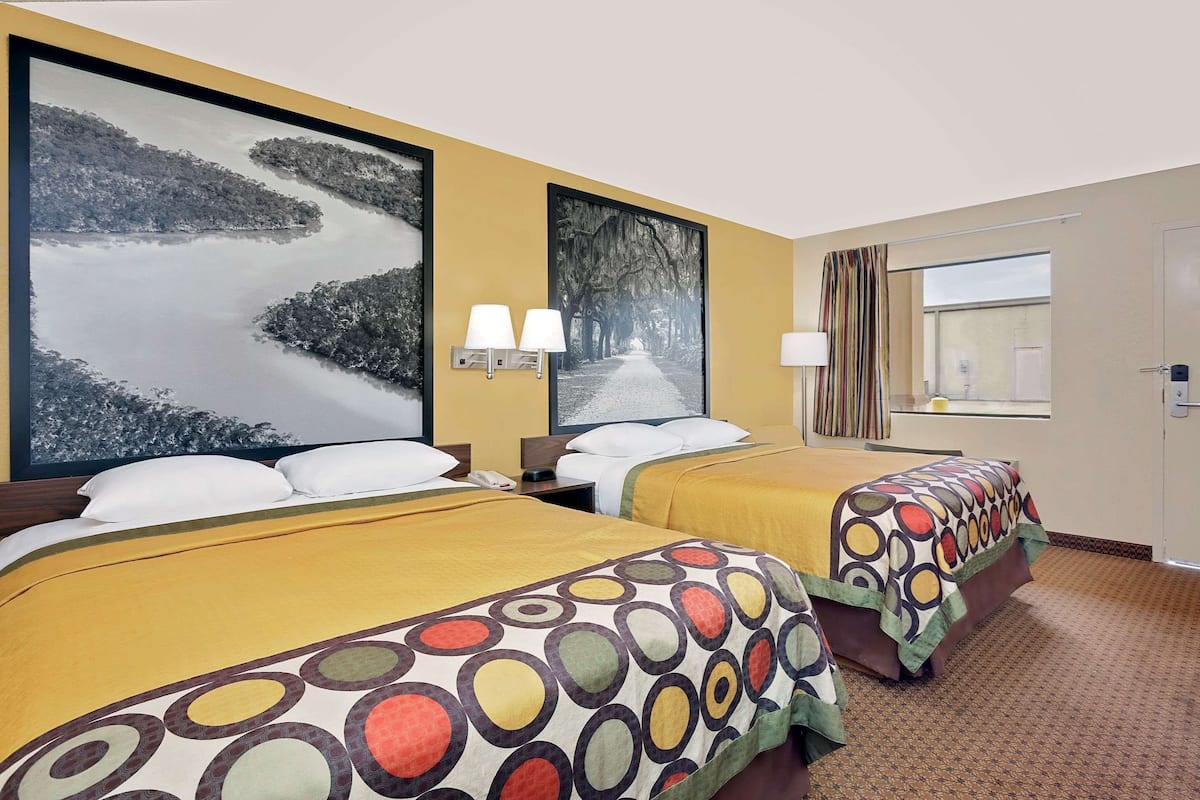 Een hotelkamer met twee bedden, elk met een geel sprei en een kleurrijk rond patroon. Twee wandlampen, een groot lijstwerk met een foto van een pad omzoomd door bomen, en een raam met gordijnen.