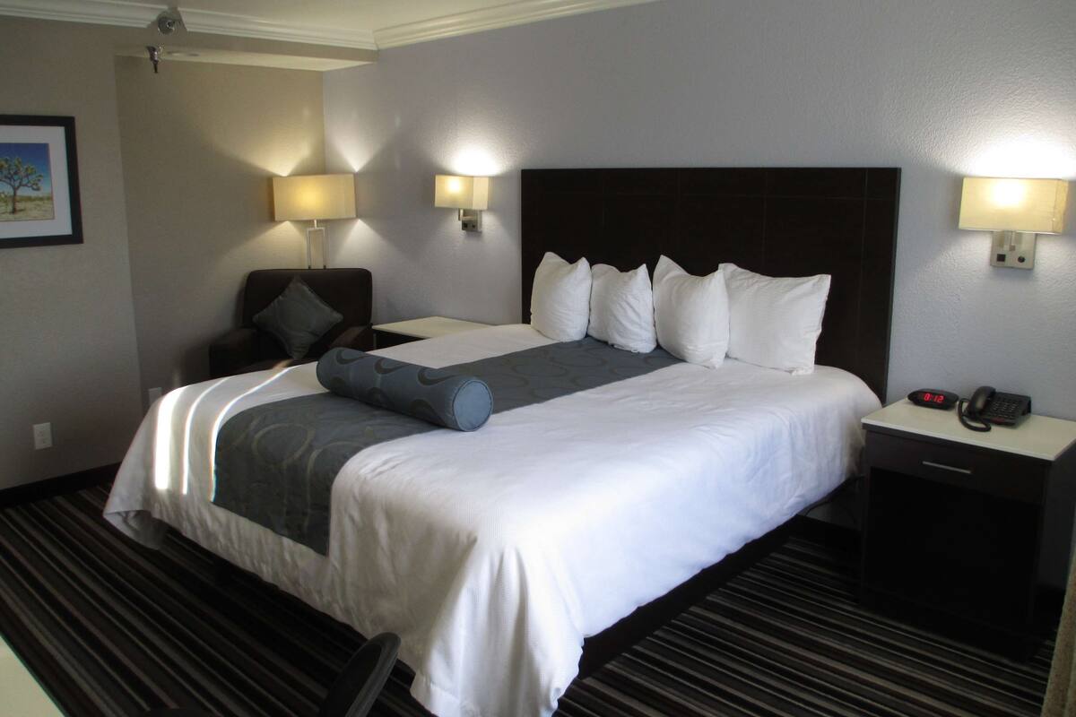Une chambre d’hôtel avec un grand lit, deux tables de chevet, une chaise et une petite table de nuit avec un téléphone et une horloge.