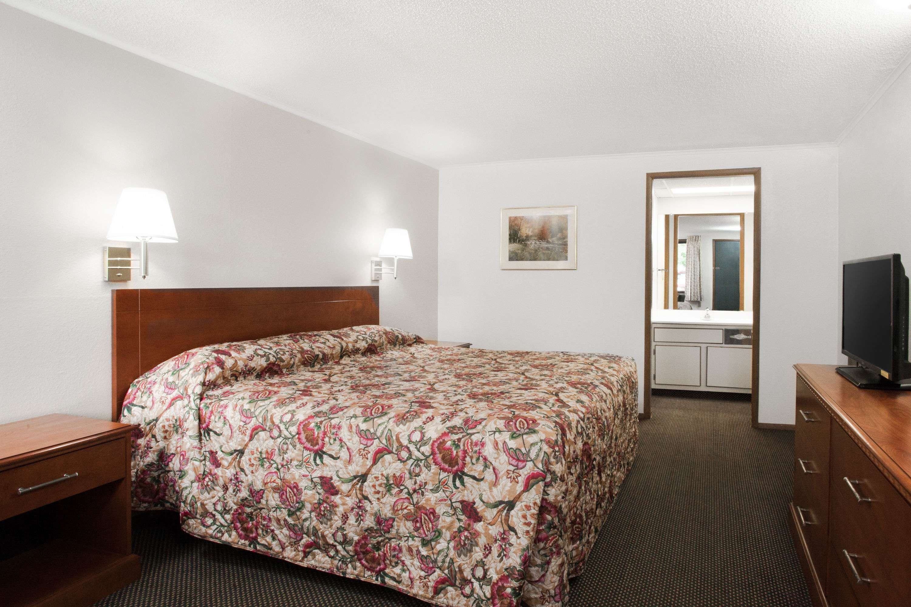 A hotel room with a floral bedspread, a wooden nightstand, a television on a wooden dresser, and a bathroom visible through an open door.