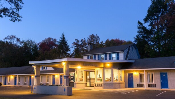 Un motel con edificio central y unidades adyacentes, que cuenta con una zona de estacionamiento techada y jardines al frente.