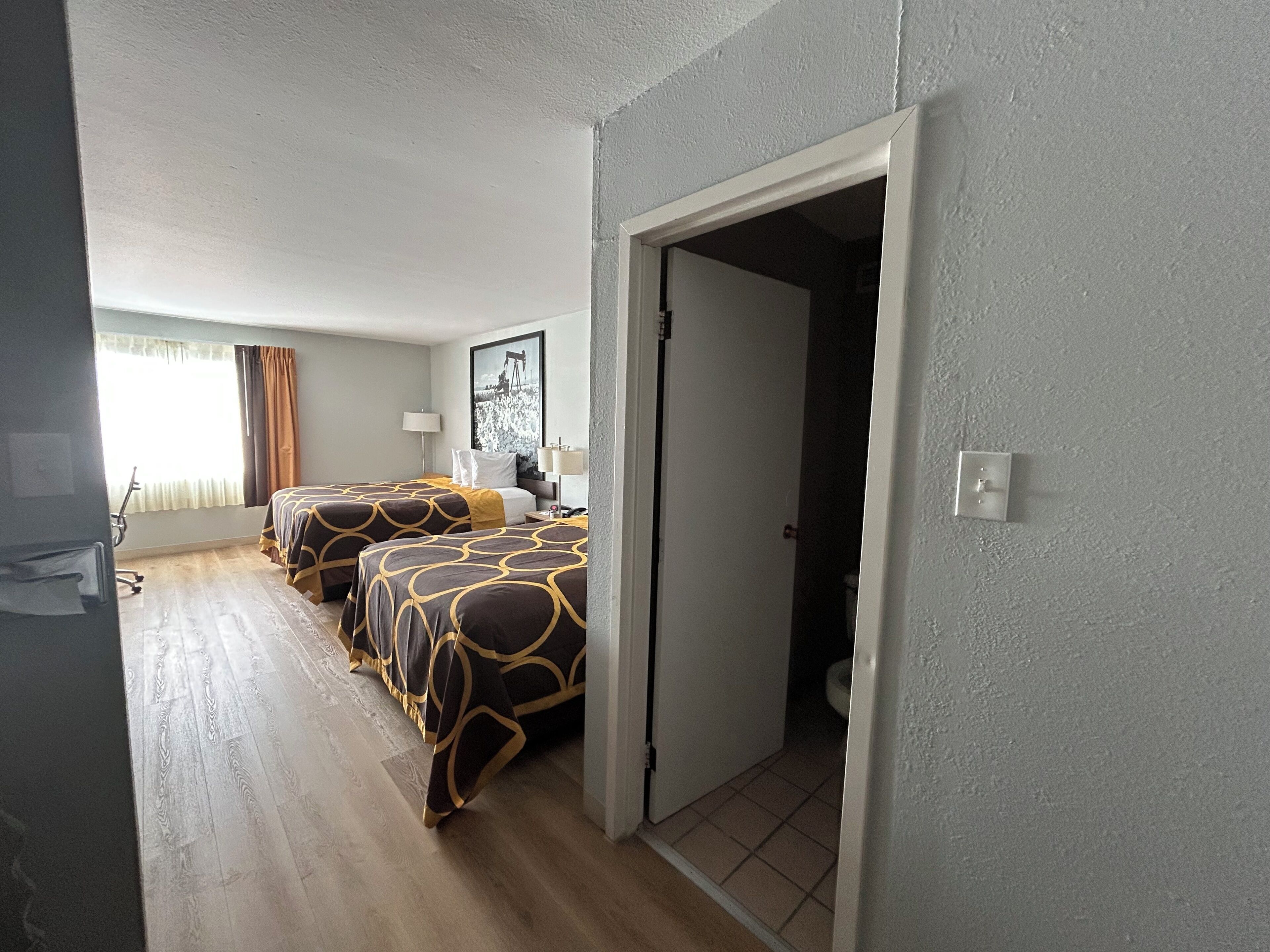 Ein Hotelzimmer mit zwei Betten, einem großen Fenster mit Vorhängen, einer Tür zu einem Badezimmer und einem gerahmten Bild an der Wand.