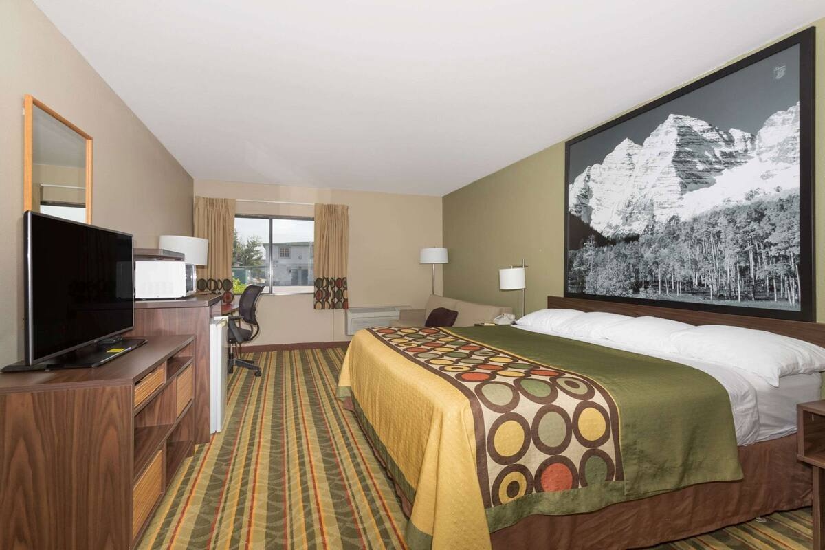 Une chambre d’hôtel avec un lit, un bureau avec une télévision, une chaise, une fenêtre et un tableau au mur.