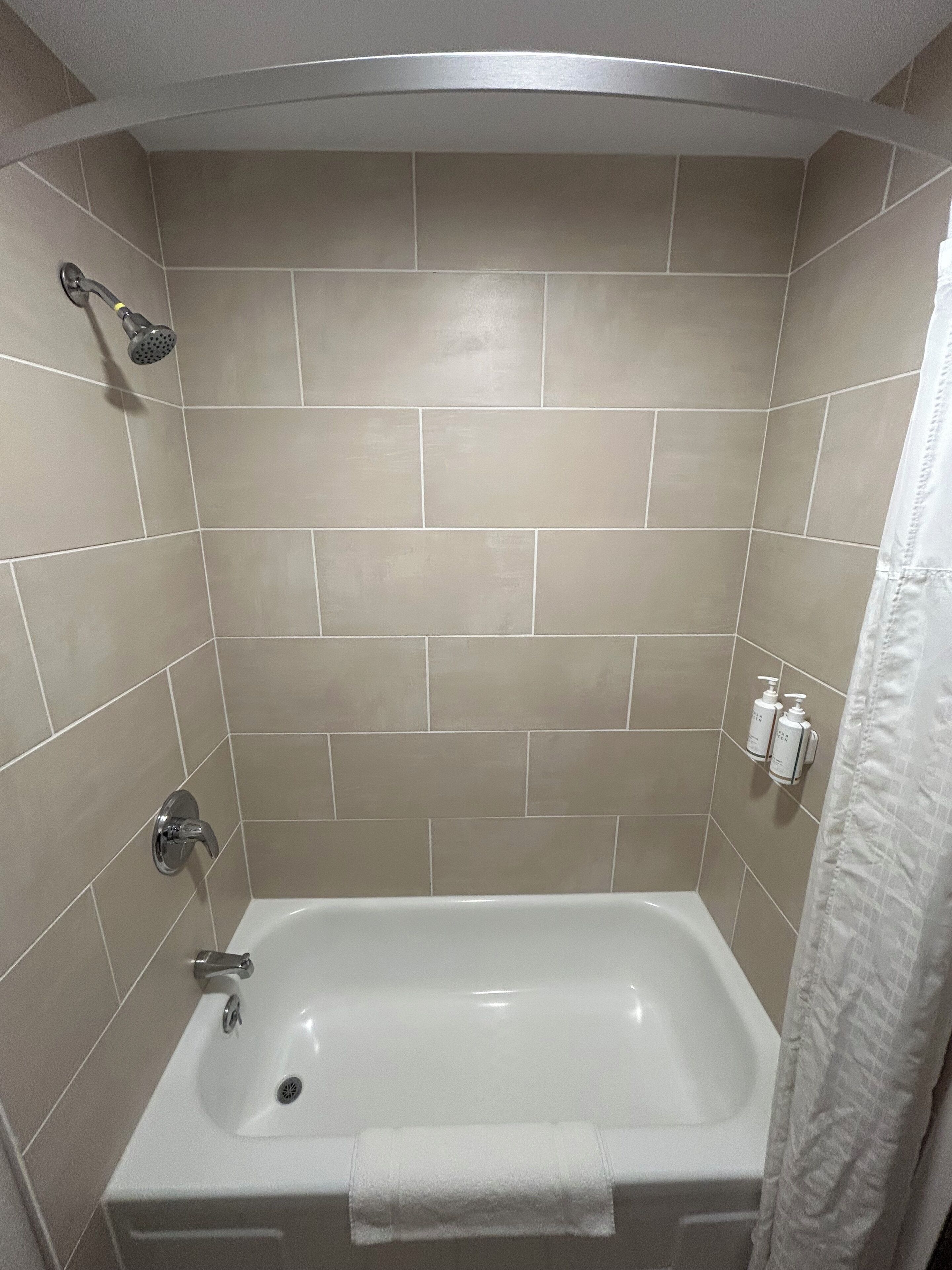 Un baño con azulejos color beige, una bañera blanca, un cabezal de ducha manual, una toalla y dos botellas de champú y acondicionador en la pared.