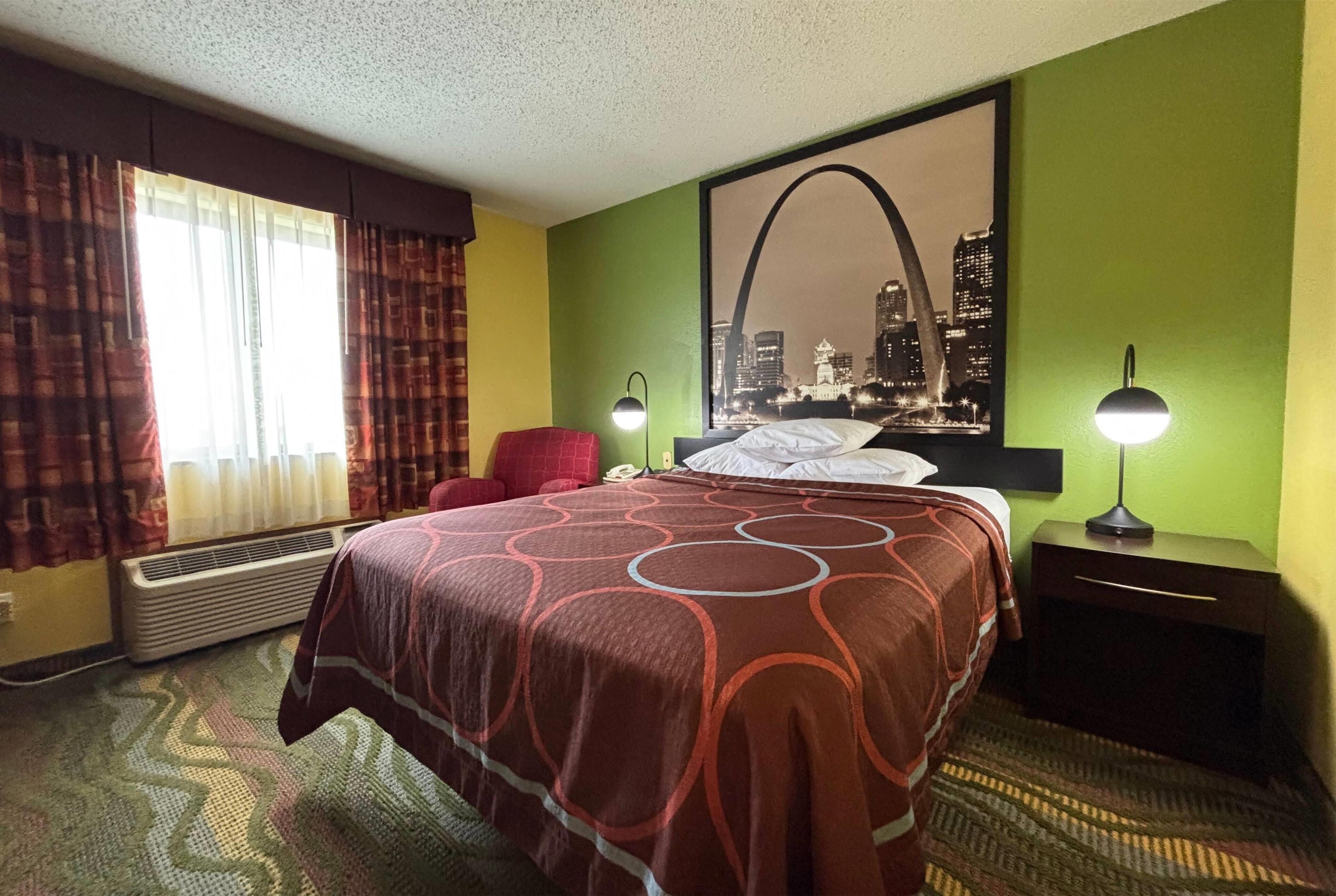 A hotel room with a large bed, a red chair, two bedside lamps, a cityscape poster on the wall, and a window with curtains.