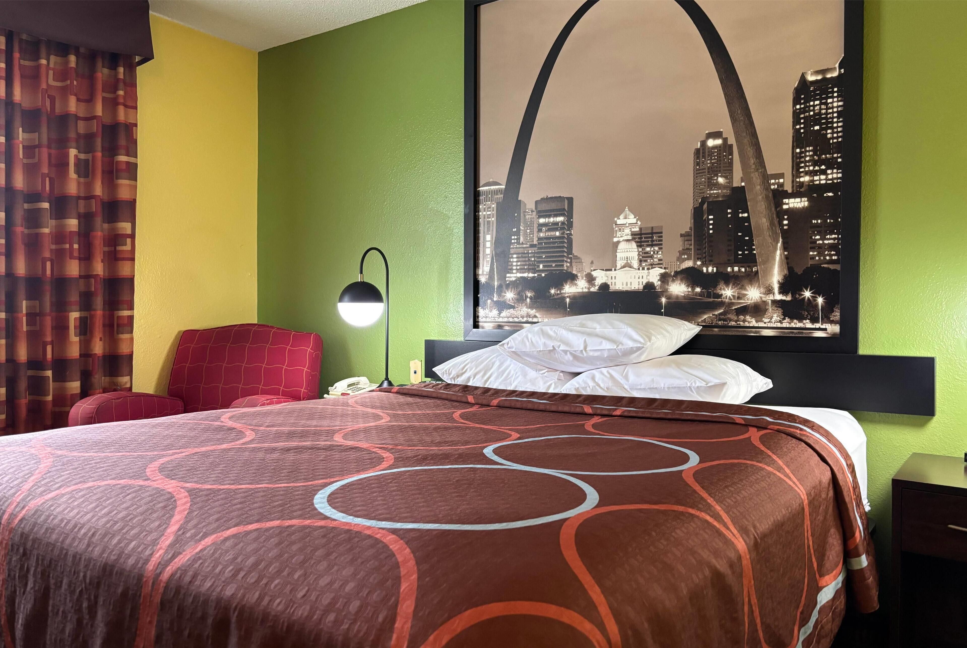 Quarto de hotel com uma cama, uma cadeira vermelha, uma mesa de cabeceira, um abajur e um pôster com vista para a cidade na parede.