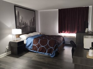 Ein Hotelzimmer mit einem Bett, einem Schreibtisch, einem Stuhl, einer Lampe, einem Fenster mit Vorhängen und einer Wand-Klimaanlage.