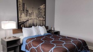 A hotel room with a bed, a nightstand, a lamp, a painting of a cityscape, and a blue and brown patterned bedspread.