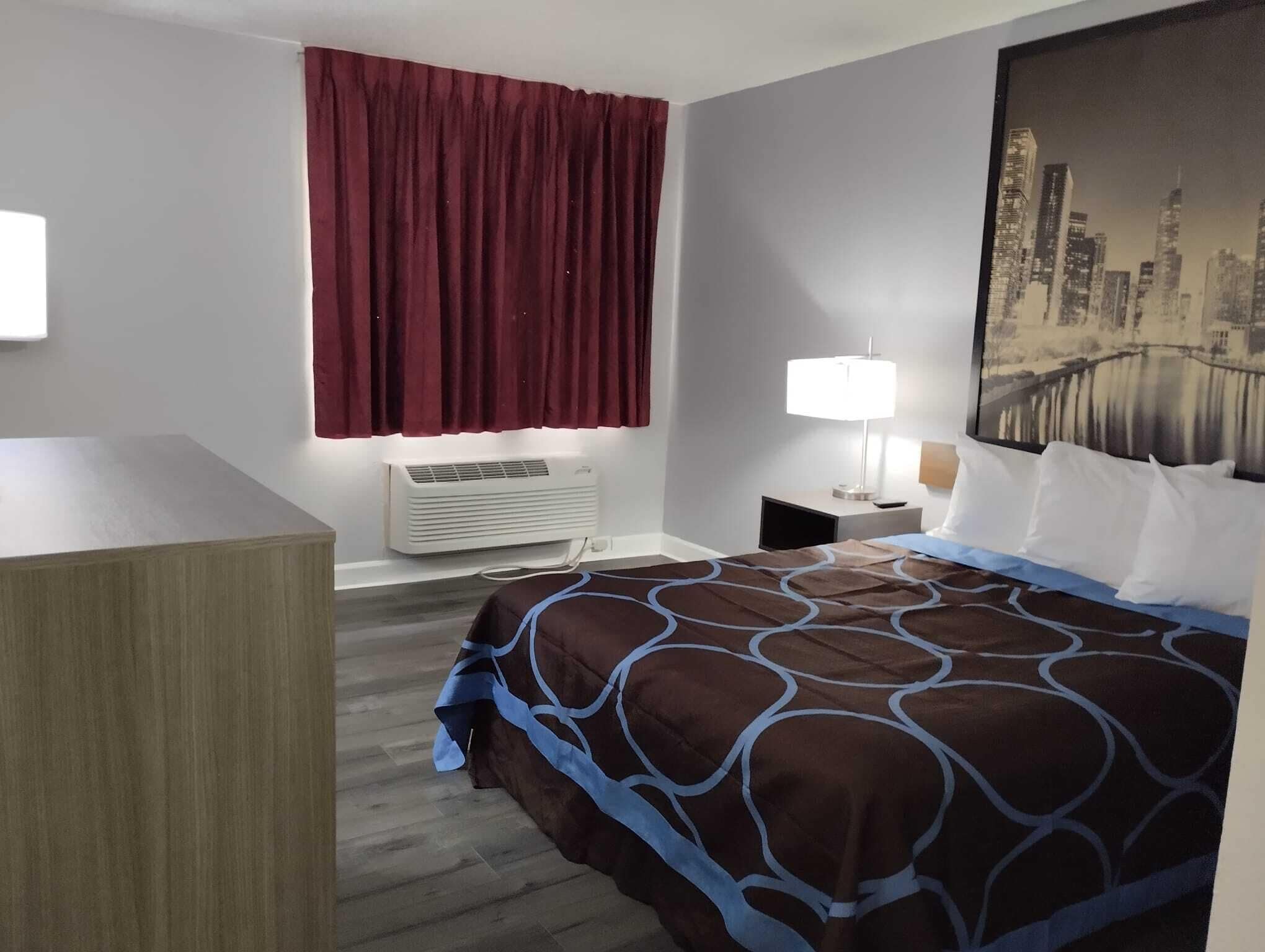 A hotel room with a bed, a desk, a window with curtains, an air conditioning unit, a nightstand with a lamp, and a cityscape painting on the wall.