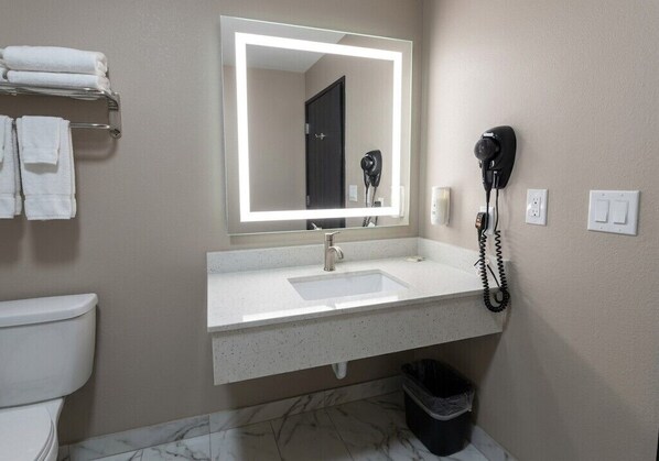 A bathroom with a white sink, a mirror with a light, a hairdryer, and a trash bin.