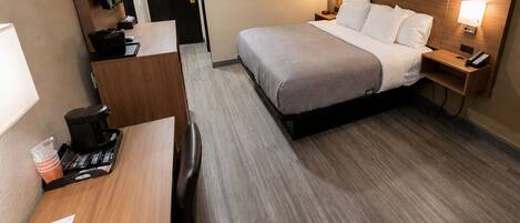 A hotel room with a bed, bedside tables, a desk, a chair, a coffee machine, and a mirror.