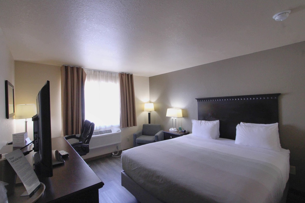 A hotel room with a large bed, a desk with a computer, two chairs, a TV, and a window with curtains.