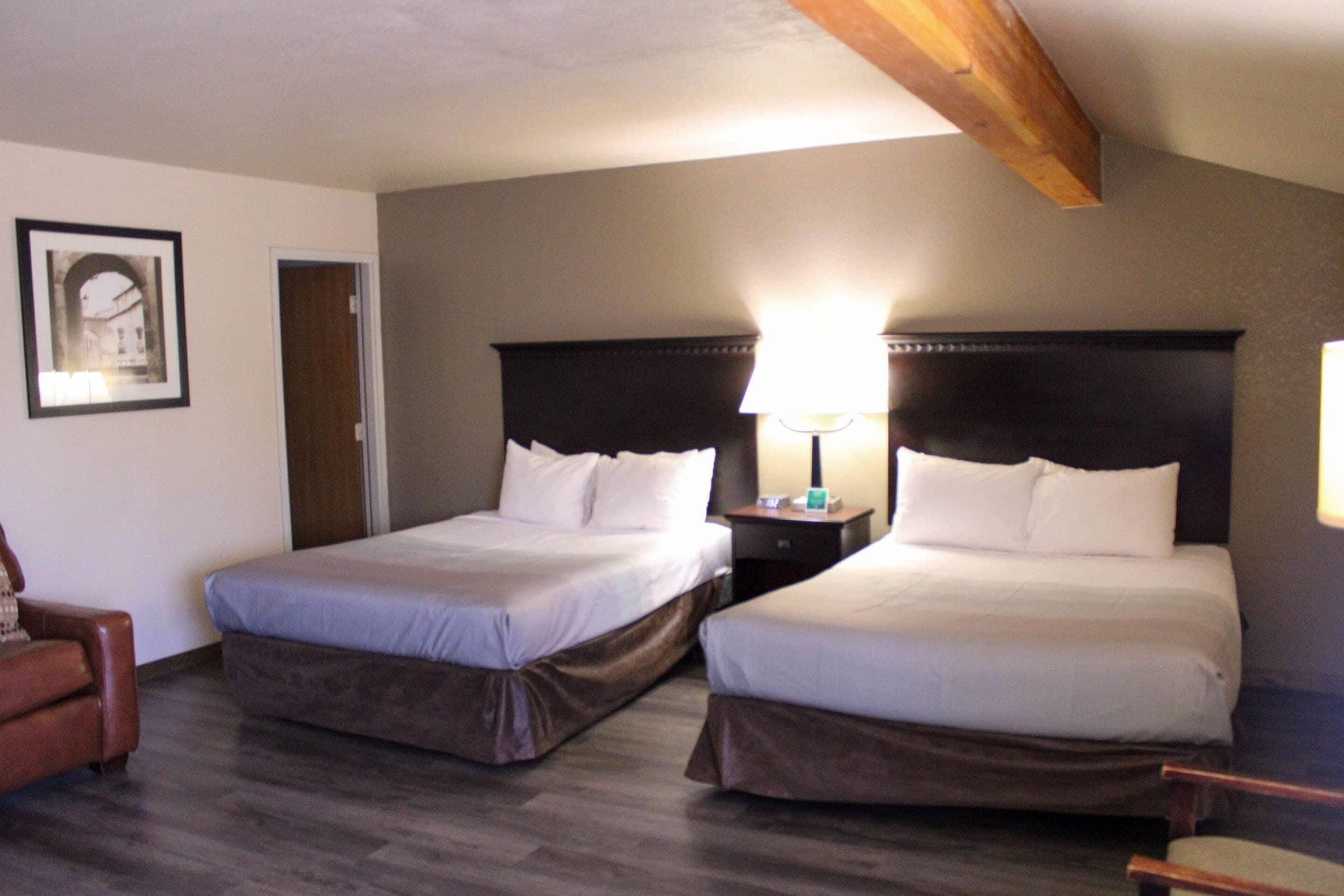A hotel room with two beds, a wooden ceiling, a framed picture on the wall, and a nightstand with a lamp.