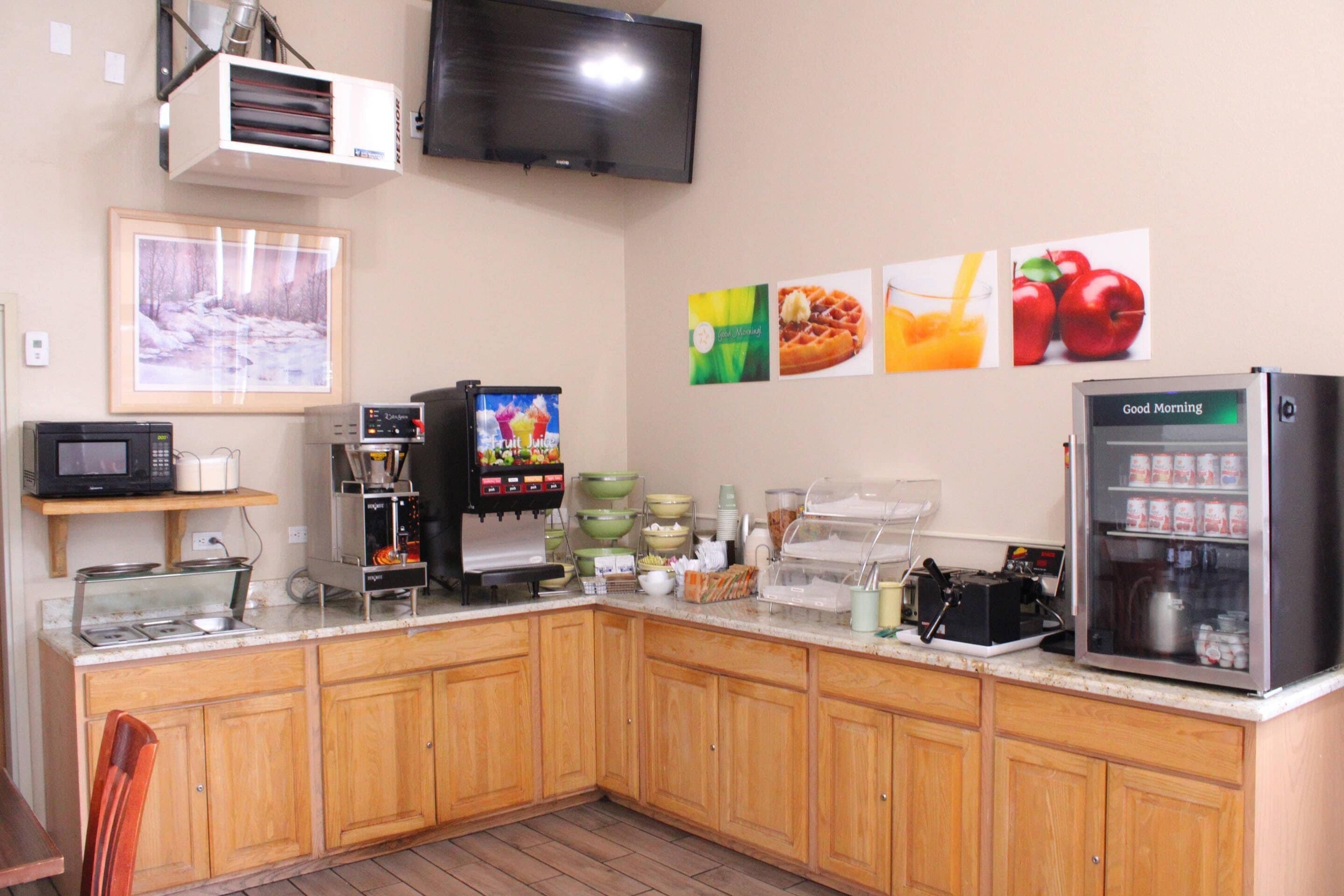 A kitchen with wooden cabinets, a microwave, a coffee machine, a refrigerator, and a television mounted on the wall.