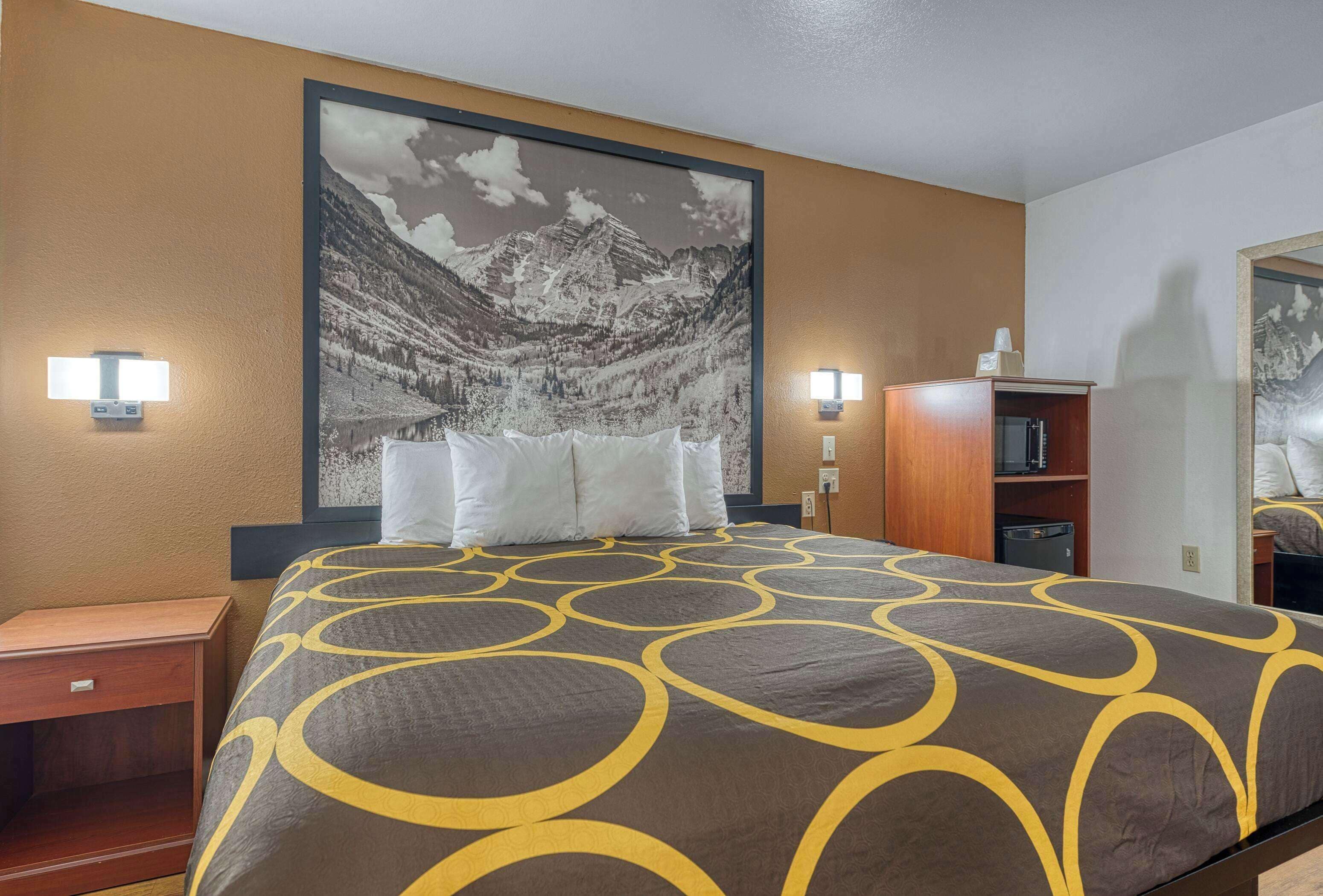 A hotel room with a large bed, two bedside lamps, a nightstand, a wooden cabinet, and a large framed picture of a mountain landscape on the wall.