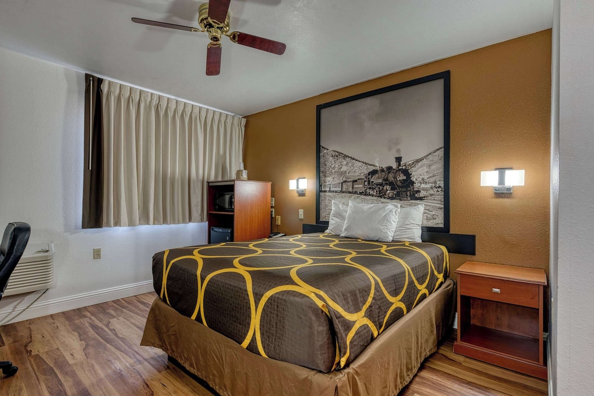 Une chambre d’hôtel équipée d’un lit, d’un bureau, d’une chaise, d’un ventilateur de plafond, d’une fenêtre avec des rideaux et d’une lampe fixée au mur.