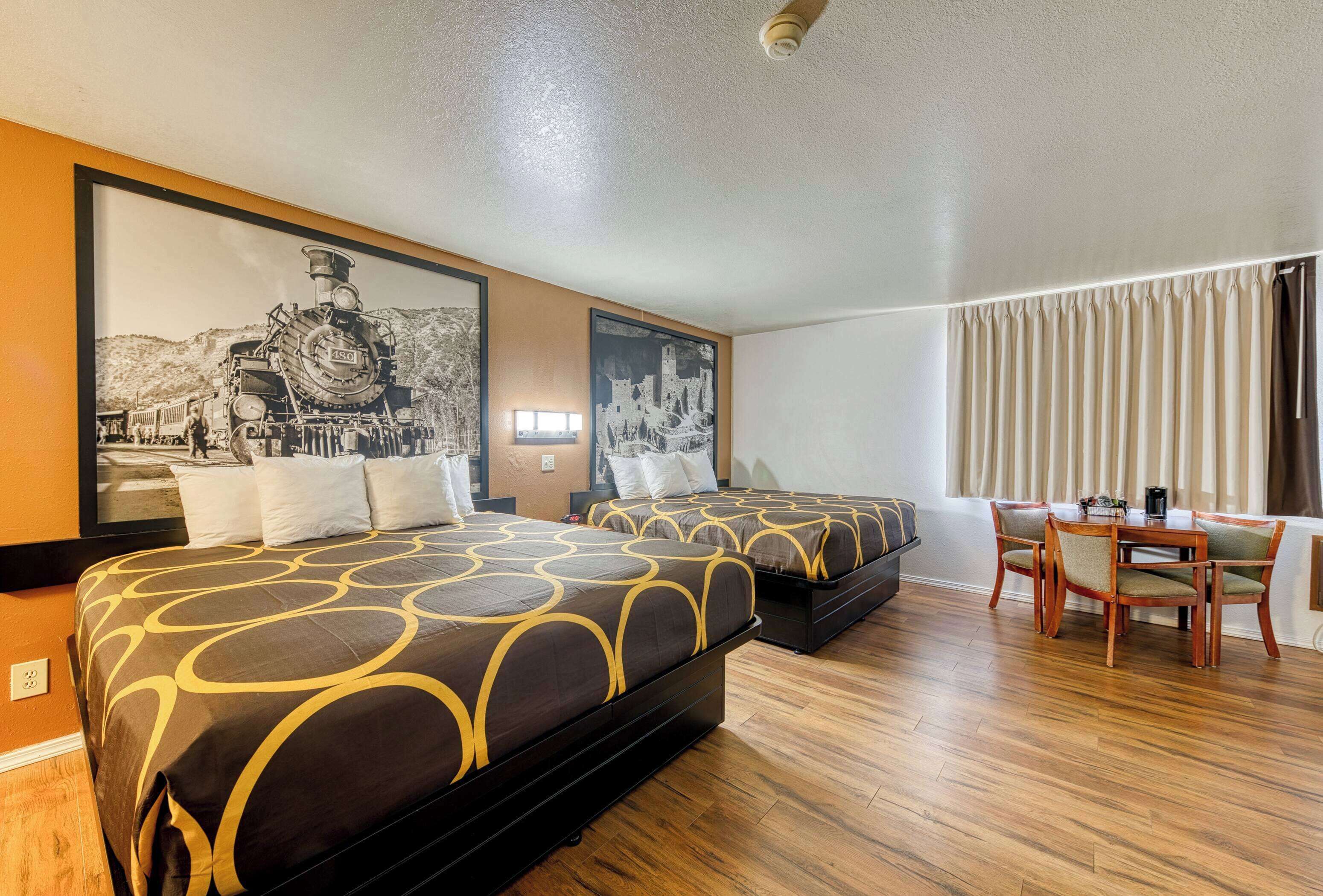 Une chambre d’hôtel avec deux lits, une table à manger avec des chaises et un grand tableau encadré représentant un train, accroché au mur.