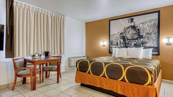 Una habitación de hotel con cama, escritorio, dos sillas, ventana con cortinas, unidad de aire acondicionado y un cuadro en la pared.