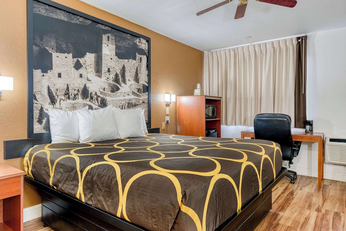 Une chambre d’hôtel avec un grand lit, un bureau, une chaise, un ventilateur de plafond et un grand tableau encadré au mur.