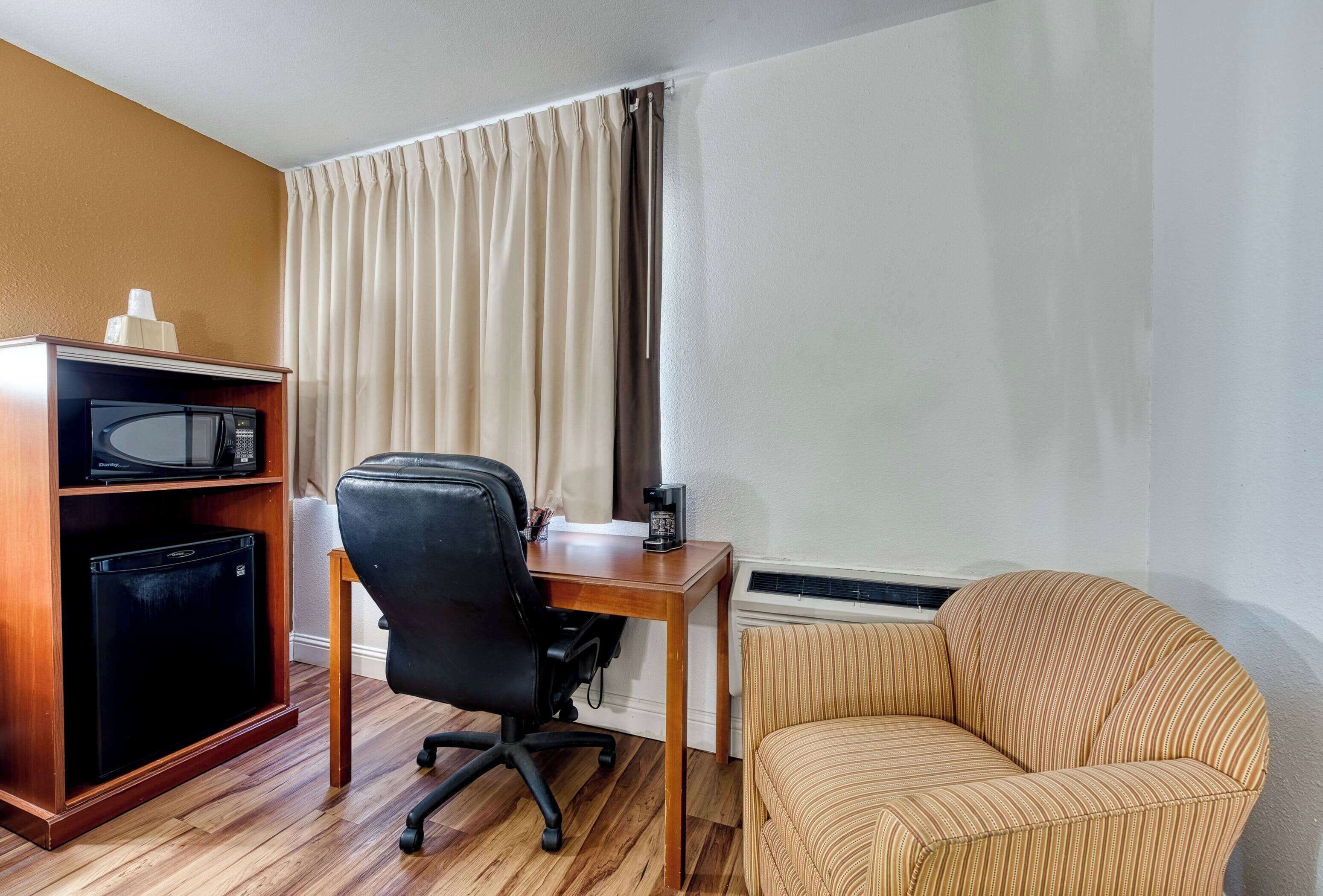 A hotel room with a wooden desk, a black office chair, a striped sofa, a minibar, and a window with curtains.